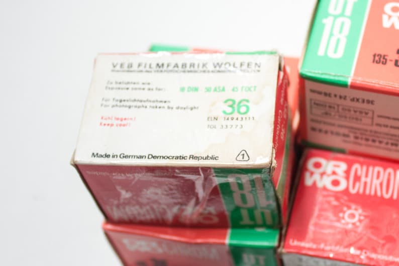 May include: A stack of three film canisters with red, white, and green labels. The top canister has the text "VEB FILMFABRIK WOLFEN" and "Made in German Democratic Republic". The middle canister has the text "OR WO CHROM" and "89 IT". The bottom canister has the text "OR WO CHROM" and "18".