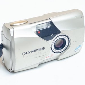 May include: A silver Olympus Stylus Epic point-and-shoot film camera featuring a 35mm f/2.8 lens, a built-in flash, and a black wrist strap with the Olympus logo. The camera body displays the Olympus brand name and Multi AF text on the front.