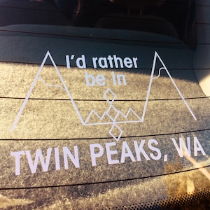May include: White decal on a car window with the text "I'd rather be in Twin Peaks, WA." The design includes stylized mountain peaks and geometric shapes, suggesting a love for the outdoors and the TV show.