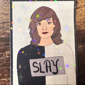 May include: A sticker with a holographic background featuring a person with brown hair and a black and white shirt. The shirt has a gray patch with the word "SLAY" written in black.