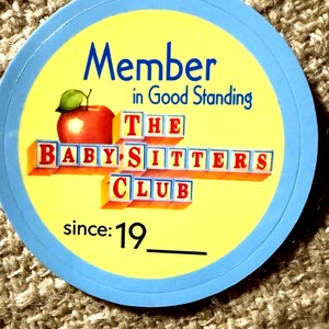 May include: A circular sticker with a light blue border and a yellow center. It features a red apple with a green leaf and the text Member in Good Standing THE BABY-SITTERS CLUB since: 19__ written in blue and red block letters.