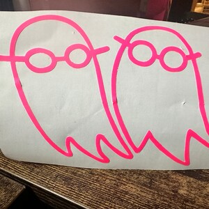 May include: Two pink ghost stickers with black outlines and pink glasses on a white background.