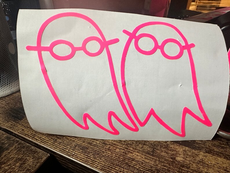 May include: Two pink ghost stickers with black outlines and pink glasses on a white background.