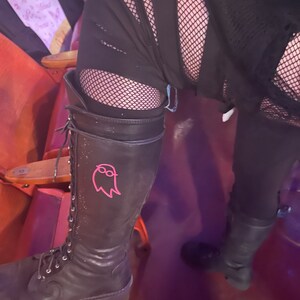 May include: Black leather lace-up boots with a pink ghost graphic on the side. The boots have a chunky heel and are worn with black fishnet stockings and ripped black pants.