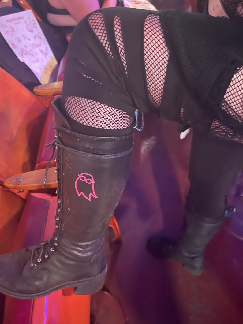 May include: Black leather lace-up boots with a pink ghost graphic on the side. The boots have a chunky heel and are worn with black fishnet stockings and ripped black pants.
