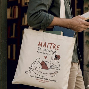 May include: A cream-colored canvas tote bag featuring a graphic illustration of a person relaxing on an inflatable pool ring. The text reads MAITRE En vacances Ne pas d&eacute;ranger ! Merci de m'avoir aid&eacute;e &agrave; grandir ! De la part de Jade 2023-2024.