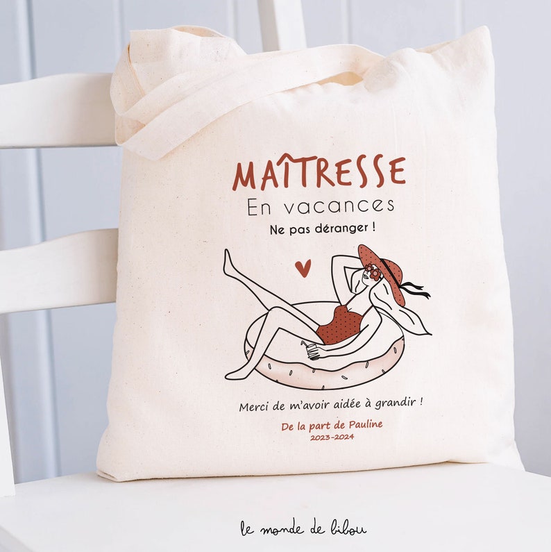 May include: A cream-colored canvas tote bag featuring a graphic of a person in a red swimsuit and hat lounging on a pool float. The text reads: MA&Icirc;TRESSE En vacances Ne pas d&eacute;ranger! Merci de m'avoir aid&eacute;e &agrave; grandir! De la part de Pauline 2023-2024.