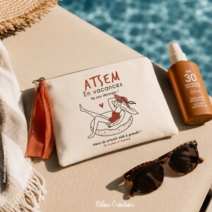 May include: A cream-colored canvas zipper pouch featuring a rust-colored tassel and a graphic illustration of a person in a red swimsuit floating on a ring. The text reads ATSEM En vacances Ne pas d&eacute;ranger! Merci de m'avoir aid&eacute; &agrave; grandir!
