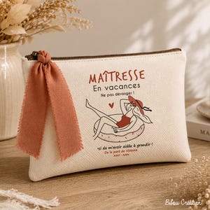 May include: A cream-colored canvas zippered pouch featuring a terracotta-colored ribbon tied to the zipper pull. The front displays a graphic of a person in a swimsuit on a float, with French text including "MA&Icirc;TRESSE En vacances" and "2023-2024".