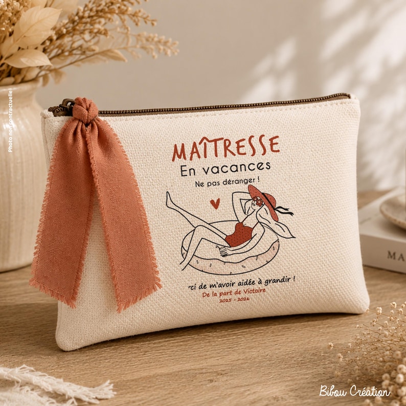 May include: A cream-colored canvas zippered pouch featuring a terracotta-colored ribbon tied to the zipper pull. The front displays a graphic of a person in a swimsuit on a float, with French text including "MA&Icirc;TRESSE En vacances" and "2023-2024".