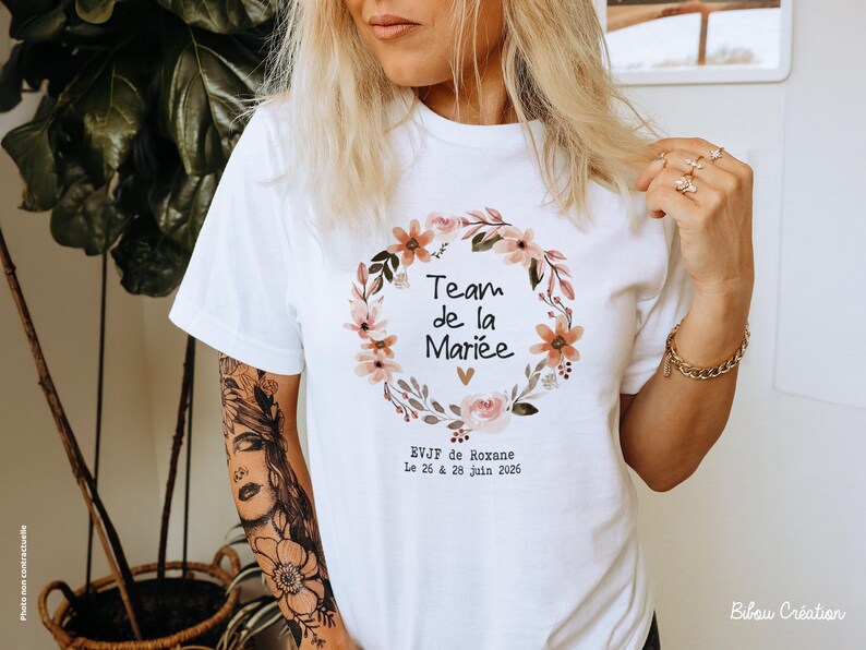 Peut inclure: T-shirt blanc avec une couronne florale et le texte "Team de la Mariée". La couronne comprend des fleurs roses et oranges. En dessous, le texte "EVJF de Roxane Le 26 & 28 Juin 2026".