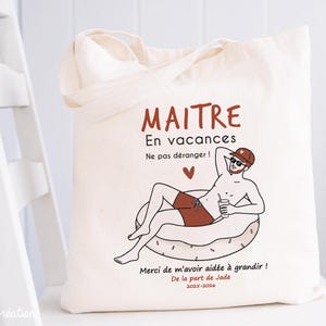 May include: A cream-colored canvas tote bag featuring a graphic illustration of a person relaxing on an inflatable pool float. The text reads MAITRE En vacances Ne pas d&eacute;ranger ! Merci de m'avoir aid&eacute;e &agrave; grandir ! De la part de Jade 2025-2026.
