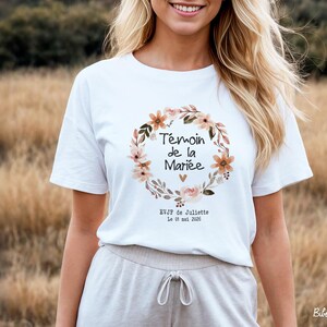 Op de afbeelding: Wit T-shirt met een bloemenkransontwerp. De krans omcirkelt de tekst "T&eacute;moin de la Mari&eacute;e" in het zwart, met extra tekst eronder. Het T-shirt heeft korte mouwen en wordt gedragen door een persoon in een veld.