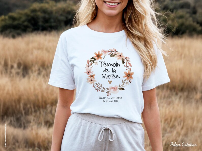Op de afbeelding: Wit T-shirt met een bloemenkransontwerp. De krans omcirkelt de tekst "T&eacute;moin de la Mari&eacute;e" in het zwart, met extra tekst eronder. Het T-shirt heeft korte mouwen en wordt gedragen door een persoon in een veld.