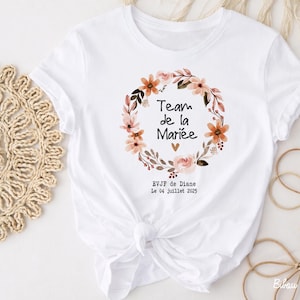 Op de afbeelding: Wit T-shirt met een bloemenkransontwerp en de tekst "Team de la Mari&eacute;e". De krans bevat perzik- en roze bloemen en groene bladeren. Extra tekst bevat "EVJF de Diane" en de datum "Le 04 Juillet 2025".