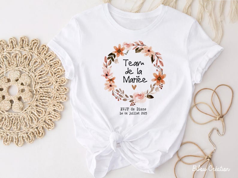 Op de afbeelding: Wit T-shirt met een bloemenkransontwerp en de tekst "Team de la Mari&eacute;e". De krans bevat perzik- en roze bloemen en groene bladeren. Extra tekst bevat "EVJF de Diane" en de datum "Le 04 Juillet 2025".
