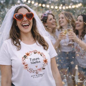 Op de afbeelding: Een vrouw met een hartvormige zonnebril en sluier lacht. Ze draagt een wit T-shirt met een bloemenkrans en de tekst "Future Mari&eacute;e". Andere vrouwen op de achtergrond houden champagneglazen vast. Confetti valt.