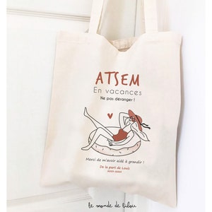 May include: A cream-colored canvas tote bag featuring a graphic illustration of a person in a swimsuit and sun hat relaxing on a pool float. The text reads ATSEM En vacances Ne pas d&eacute;ranger ! Merci de m'avoir aid&eacute; &agrave; grandir ! De la part de Louis 2023-2024.
