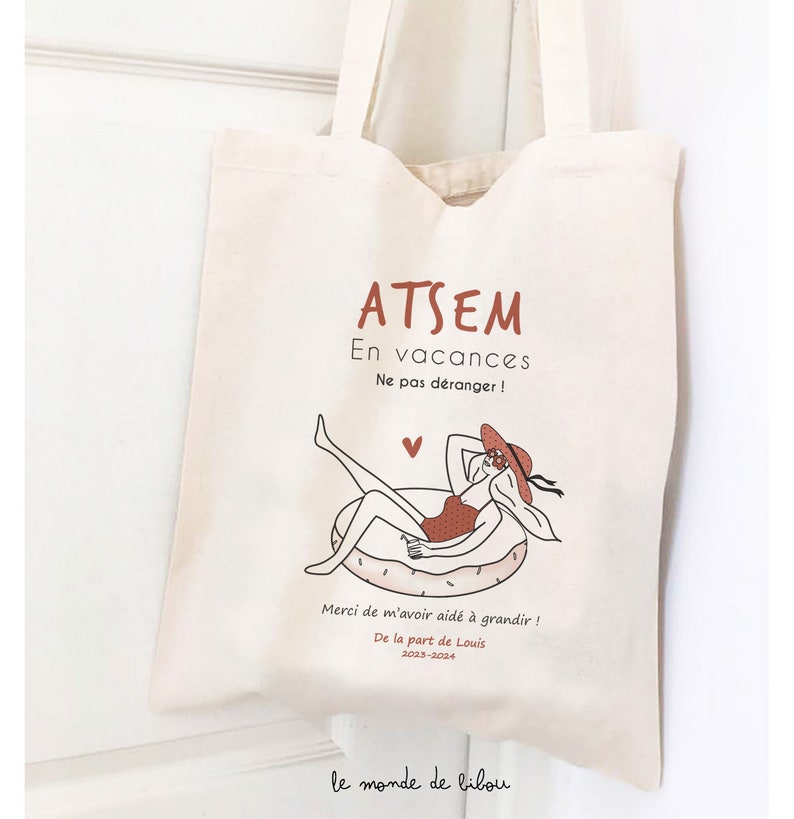 May include: A cream-colored canvas tote bag featuring a graphic illustration of a person in a swimsuit and sun hat relaxing on a pool float. The text reads ATSEM En vacances Ne pas d&eacute;ranger ! Merci de m'avoir aid&eacute; &agrave; grandir ! De la part de Louis 2023-2024.