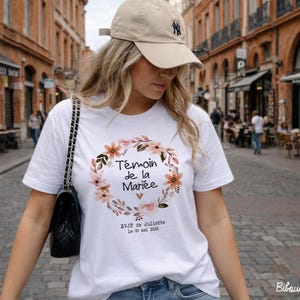 Op de afbeelding: Wit T-shirt met een bloemenkransontwerp en de tekst "T&eacute;moin de la Mari&eacute;e". Het shirt bevat ook de tekst "EVJF de Juliette Le 01 mai 2020". De persoon draagt een beige baseballpet en een zwarte handtas.