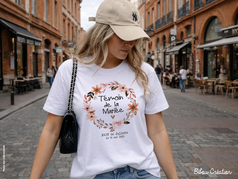 Op de afbeelding: Wit T-shirt met een bloemenkransontwerp en de tekst "T&eacute;moin de la Mari&eacute;e". Het shirt bevat ook de tekst "EVJF de Juliette Le 01 mai 2020". De persoon draagt een beige baseballpet en een zwarte handtas.