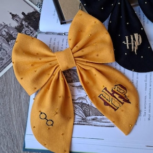 Puede incluir: Lazo de tela amarillo mostaza con detalles de cuentas doradas y bordados de gafas y el logo "HP". También se ve un lazo negro con cuentas doradas y el logo "HP". Los lazos están sobre un libro con texto.