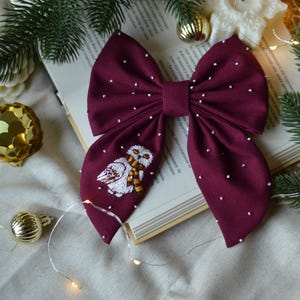 May include: A burgundy bow with white polka dots and an embroidered white owl with a brown scarf. The bow is on a book with a white background.