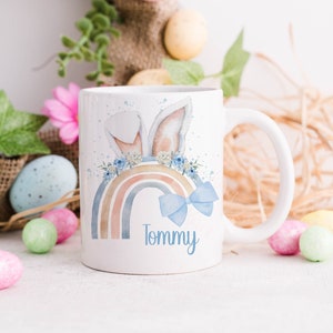 May include: White ceramic mug with a rainbow, bunny ears, and a blue bow. The mug has the name "Tommy" written on it.