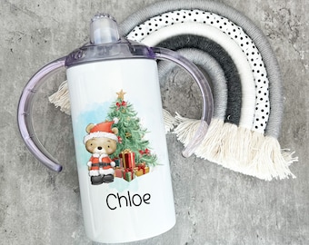 Pode incluir: Um copo de aço inoxidável branco com um design de Natal que apresenta um urso usando um chapéu de Papai Noel e uma árvore de Natal. O copo tem duas alças de plástico transparente e o nome "Chloe" está impresso na frente.
