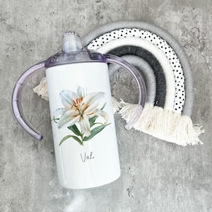 Puede incluir: Un vaso de acero inoxidable blanco con un dise&ntilde;o de flor de lirio blanco y el nombre "Val" impreso en la parte delantera. El vaso tiene dos asas y una tapa morada.