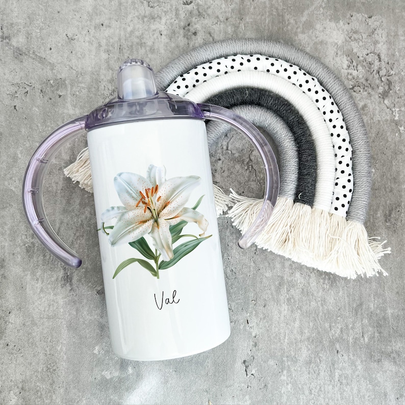 Puede incluir: Un vaso de acero inoxidable blanco con un dise&ntilde;o de flor de lirio blanco y el nombre "Val" impreso en la parte delantera. El vaso tiene dos asas y una tapa morada.