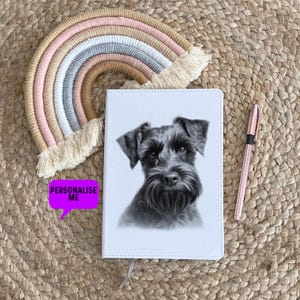 Pode incluir: Um diário de couro branco com uma ilustração em preto e branco de um schnauzer na capa. O diário está sobre um tapete tecido com um macramê arco-íris pendurado acima.