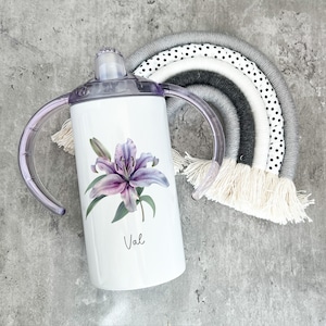 Puede incluir: Un vaso de acero inoxidable blanco con un dise&ntilde;o de lirio morado y el nombre "Val" impreso en la parte delantera. El vaso tiene dos asas y una tapa morada.