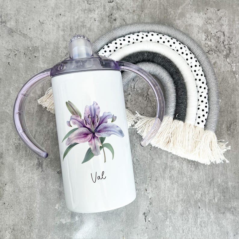 Puede incluir: Un vaso de acero inoxidable blanco con un dise&ntilde;o de lirio morado y el nombre "Val" impreso en la parte delantera. El vaso tiene dos asas y una tapa morada.