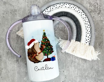 Pode incluir: Uma caneca de treinamento de aço inoxidável branco com uma alça e uma tampa de plástico transparente. A caneca apresenta um ouriço de desenho animado decorando uma árvore de Natal com o nome "Caitlin" impresso abaixo da imagem.
