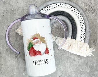 Pode incluir: Um copo branco de aço inoxidável com uma ilustração do Papai Noel e o nome "THOMAS" impresso na frente. O copo tem duas alças de plástico transparente e está ao lado de um arco-íris cinza e branco.