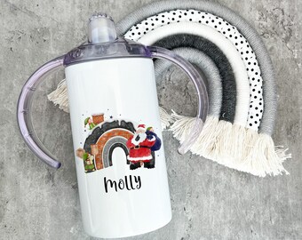 Pode incluir: Copo de aço inoxidável branco com uma cena de Natal com Papai Noel, duendes e um arco-íris. O copo tem duas alças e o nome "molly" impresso na frente.