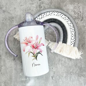 Puede incluir: Un vaso de acero inoxidable blanco con dos asas, con un dise&ntilde;o de flor de lirio rosa y la palabra "Nana".