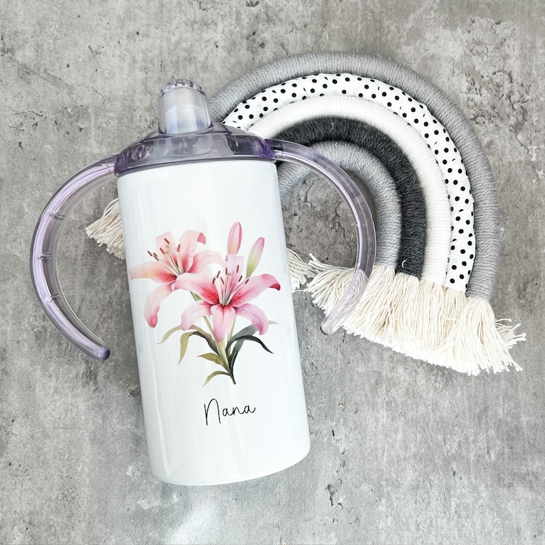 Puede incluir: Un vaso de acero inoxidable blanco con dos asas, con un dise&ntilde;o de flor de lirio rosa y la palabra "Nana".