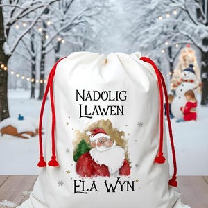 Puede incluir: Una bolsa de regalo de Navidad blanca con cordones de extracción rojos. La bolsa presenta una ilustración de acuarela de Santa Claus con el texto "Nadolig Llawen Ela Wyn".