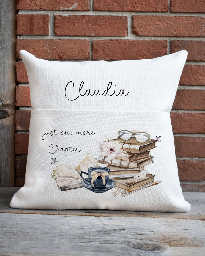Peut inclure: Un coussin carr&eacute; blanc avec une poche frontale portant le pr&eacute;nom Claudia en &eacute;criture cursive noire. Le motif repr&eacute;sente une pile de livres, une tasse &agrave; th&eacute; bleu fonc&eacute;, des lunettes et le texte just one more Chapter en lettres noires.