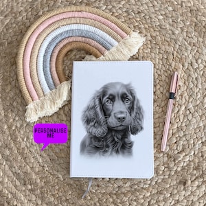 Pode incluir: Um diário de couro branco com um desenho a lápis preto e branco de um filhote de cocker spaniel preto na capa. O diário está sobre um tapete tecido com um macramé arco-íris pendurado acima.