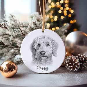 May include: A white, circular ceramic Christmas ornament with a detailed pencil sketch of a dog's face. The dog's name, "Peggy," is written in cursive below the portrait. It hangs from a gold ribbon, suitable for festive decorations.