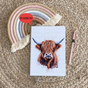 Pode incluir: Um caderno branco com uma ilustração de aquarela de uma vaca das terras altas na capa. O caderno tem uma borla cinza e as palavras "Personalize-me" estão escritas em vermelho em um balão de fala acima do caderno.