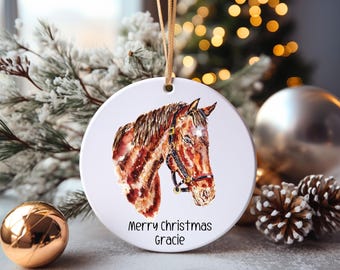 May include: A round white ceramic ornament with a watercolor painting of a brown horse's head. The ornament has a gold string for hanging and the text "Merry Christmas Gracie" printed on the bottom.