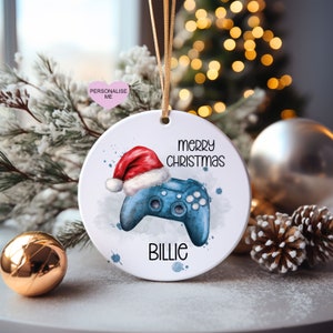May include: A white ceramic Christmas bauble featuring a blue video game controller wearing a red Santa hat. The bauble is inscribed with the text "merry christmas" and "Billie".