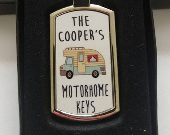Puede incluir: Un llavero plateado con una etiqueta rectangular blanca. La etiqueta tiene una ilustración de dibujos animados de una autocaravana y el texto "The Cooper's Motorhome Keys".