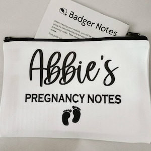 May include: White zippered pouch with black text that reads "Abbie's Pregnancy Notes" and a pair of black footprints.