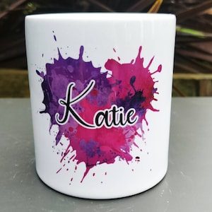 May include: White ceramic mug with a pink and purple watercolour heart design and the name "Katie" in black script.