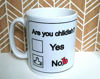 Könnte beinhalten: Weiße Keramiktasse mit schwarzem Rand und dem Text "Are you childish?" mit zwei Kontrollkästchen. Das obere Kontrollkästchen ist leer und der Text "Yes" befindet sich darunter. Das untere Kontrollkästchen enthält eine Zeichnung eines Mittelfingers und den Text "No" mit einem roten Herzen daneben.